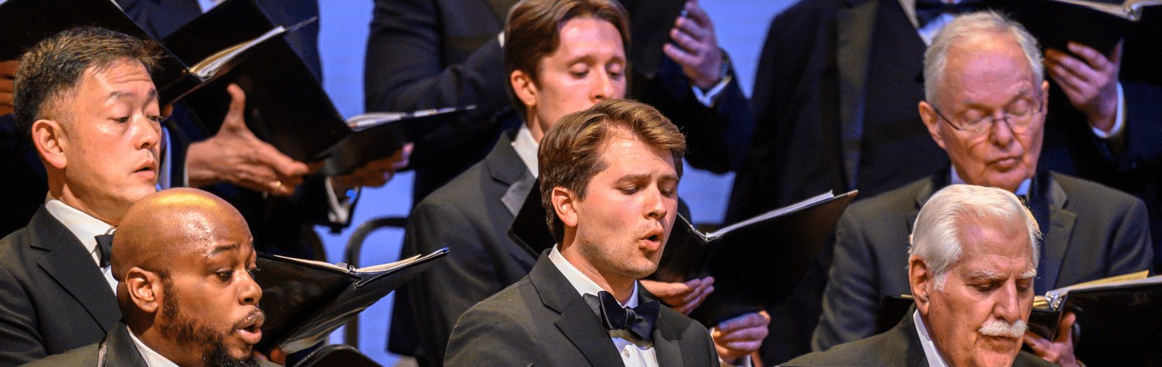 A group of men on a stage, singing, with one man in front conducting them.