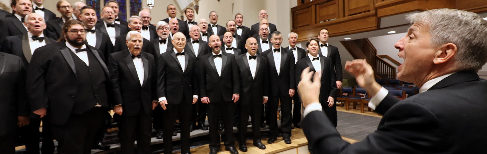 A group of men on a stage, singing, with one man in front conducting them.